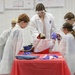 A science educator in a white lab coat and safety goggles leads a hands-on science demonstration for a group of children in a classroom setting. All participants are wearing white lab coats and protective safety goggles. The activity takes place around a red-tablecloth-covered table with various science materials and props.
