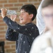 A young boy, approximately 10-12 years old, is laughing and holding a homemade device constructed from a cardboard tube wrapped in aluminum foil, likely a DIY telescope or solar viewer. He appears to be in a school or educational setting, indicated by the brick wall background. The scene suggests a hands-on science or STEM activity, possibly related to solar observation.