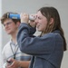 A young woman in a gray fleece jacket is looking through a homemade tube viewer, likely a DIY solar viewer or pinhole viewer constructed from a cardboard tube wrapped with blue tape. She appears amused or excited, squinting one eye while peering through the device. A young man wearing glasses and a striped shirt stands blurred in the background.