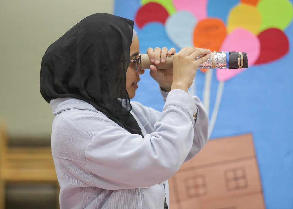 A young girl wearing a black hijab and glasses is looking through a homemade telescope or periscope constructed from cardboard tubes and foil. She is standing in what appears to be a classroom or library setting. The background features a colorful bulletin board display with balloons and a house, consistent with an educational environment.