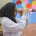 A young girl wearing a black hijab and glasses is looking through a homemade telescope or periscope constructed from cardboard tubes and foil. She is standing in what appears to be a classroom or library setting. The background features a colorful bulletin board display with balloons and a house, consistent with an educational environment.
