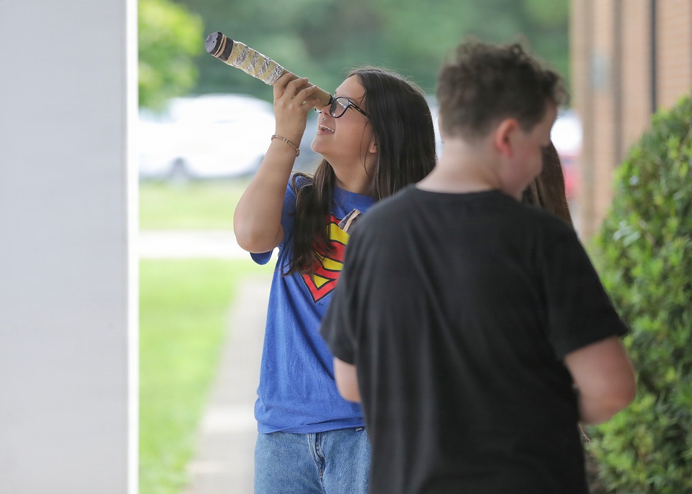A young girl wearing glasses and a Superman t-shirt is looking through a homemade telescope or tube-shaped viewing device while standing outdoors. A second person in a black t-shirt stands nearby with their back to the camera. The setting appears to be outside a brick building with green landscaping.