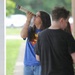 A young girl wearing glasses and a Superman t-shirt is looking through a homemade telescope or tube-shaped viewing device while standing outdoors. A second person in a black t-shirt stands nearby with their back to the camera. The setting appears to be outside a brick building with green landscaping.