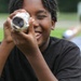 A young boy is smiling broadly while looking through a homemade solar viewer or pinhole viewer wrapped in aluminum foil. The device appears to be a cardboard tube covered in foil with a dark filter or pinhole at the end. A second child is visible in the blurred background holding a similar device, suggesting a group activity, likely related to solar eclipse viewing or a science project.