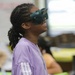 A young person wearing dark green safety goggles or a VR headset is smiling broadly while looking upward, suggesting an engaging or exciting experience. They are wearing a purple Adidas track jacket and have braided hair pulled back. The setting appears to be a classroom or educational environment based on the blurred background.