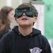 A young boy wearing dark safety goggles looks upward with an expression of curiosity or wonder. He is wearing a dark navy hoodie with a 'Flying Ace' Snoopy graphic on the chest. The setting appears to be an indoor event or fair, with other children visible in the blurred background.