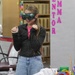 A young girl wearing VR goggles stands in what appears to be a school library or classroom, smiling and reacting with raised fists to a virtual reality experience. Behind her is a display board with pink letters spelling 'EMMA' and 'ENTOR' (likely 'MENTOR'). A table in the foreground holds art supplies including Crayola markers, scissors, and other craft materials.