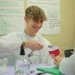 A smiling teenage male student wearing a white lab coat/apron is conducting a science experiment at a table, using a small measuring spoon over a red Solo cup. He appears to be in a classroom or educational setting with handwritten posters on the wall behind him. A second student in a lab coat is partially visible on the right side of the frame.