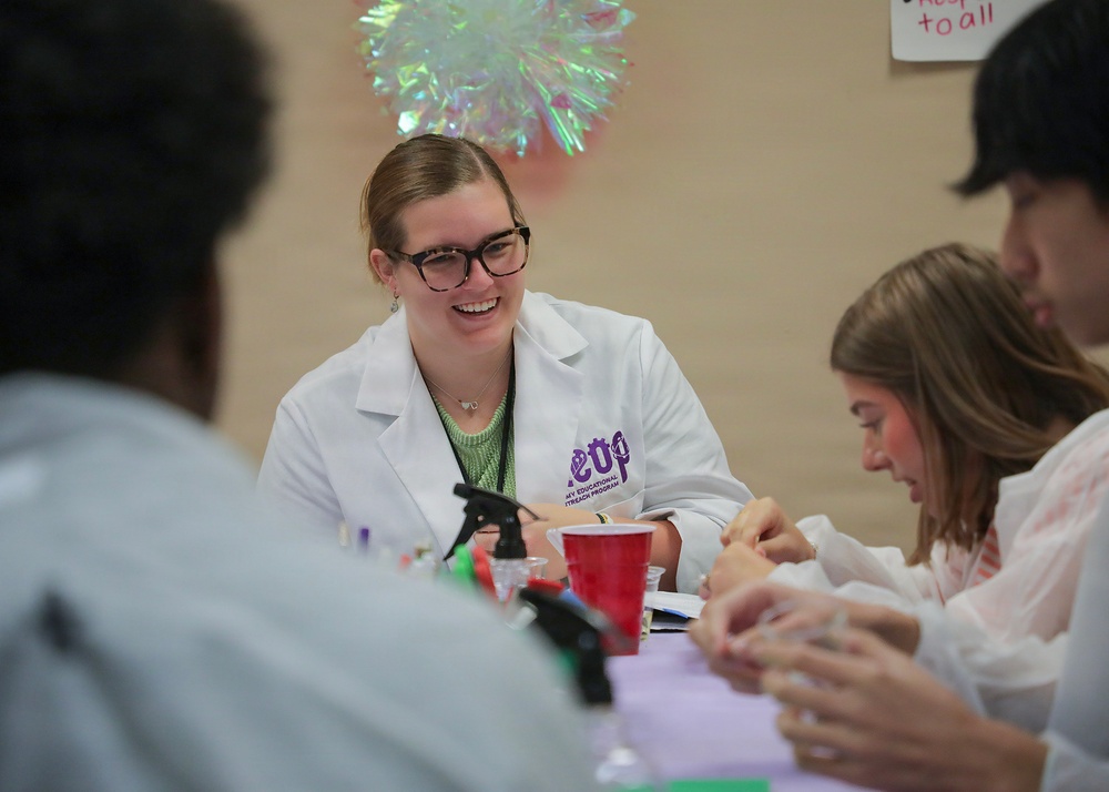 A smiling young woman wearing a white lab coat with a purple logo reading 'SEOP' (Science/Chemistry Educational Outreach Program) is seated at a table engaged in a hands-on activity with other participants. The setting appears to be an educational outreach or science demonstration event, with supplies including red cups, spray bottles, and small containers on the table. Several other participants are visible around the table, focused on the activity.