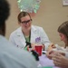 A smiling young woman wearing a white lab coat with a purple logo reading 'SEOP' (Science/Chemistry Educational Outreach Program) is seated at a table engaged in a hands-on activity with other participants. The setting appears to be an educational outreach or science demonstration event, with supplies including red cups, spray bottles, and small containers on the table. Several other participants are visible around the table, focused on the activity.