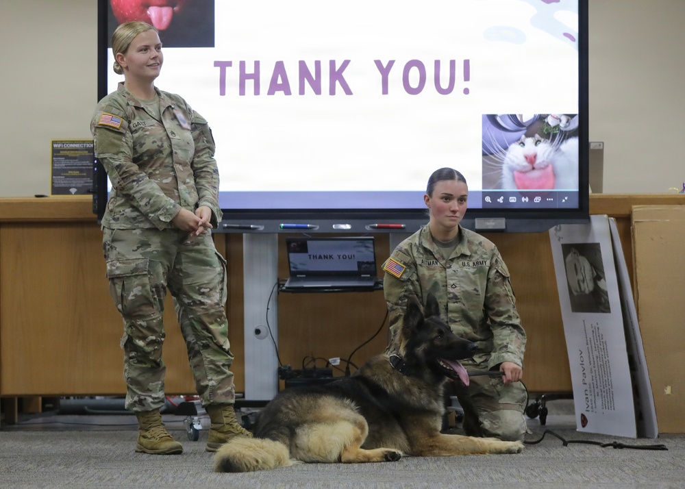 Two U.S. Army soldiers are presenting at what appears to be a military briefing or educational event, with a German Shepherd military working dog lying on the floor between them. A large display screen behind them shows a 'THANK YOU!' slide, suggesting the conclusion of a presentation. A poster to the right appears to reference 'Ivan Pavlov,' possibly related to the dog's training or history.