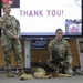 Two U.S. Army soldiers are presenting at what appears to be a military briefing or educational event, with a German Shepherd military working dog lying on the floor between them. A large display screen behind them shows a 'THANK YOU!' slide, suggesting the conclusion of a presentation. A poster to the right appears to reference 'Ivan Pavlov,' possibly related to the dog's training or history.
