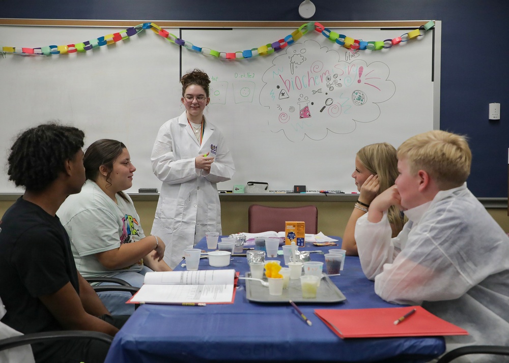 A young woman in a white lab coat stands at the front of a classroom near a whiteboard that reads 'biochemistry,' presenting to four seated students around a blue-covered table. The setting appears to be an educational workshop or science camp, with lab materials and supplies visible on the table. A colorful paper chain garland decorates the top of the whiteboard.