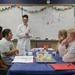 A young woman in a white lab coat stands at the front of a classroom near a whiteboard that reads 'biochemistry,' presenting to four seated students around a blue-covered table. The setting appears to be an educational workshop or science camp, with lab materials and supplies visible on the table. A colorful paper chain garland decorates the top of the whiteboard.
