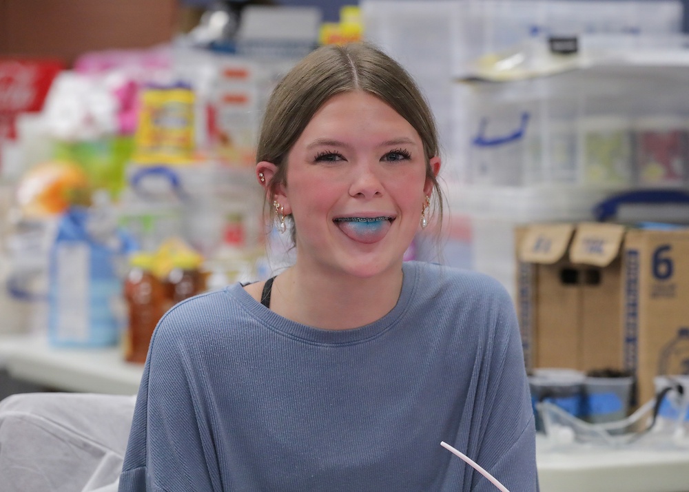 A young woman is smiling and sticking out her tongue, which appears to be stained blue, likely from food or a colored beverage. She is wearing a light blue waffle-knit sweater and small hoop earrings, and has braces visible on her teeth. The background shows a cluttered retail or storage environment with various packaged goods and boxes.