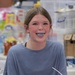 A young woman is smiling and sticking out her tongue, which appears to be stained blue, likely from food or a colored beverage. She is wearing a light blue waffle-knit sweater and small hoop earrings, and has braces visible on her teeth. The background shows a cluttered retail or storage environment with various packaged goods and boxes.