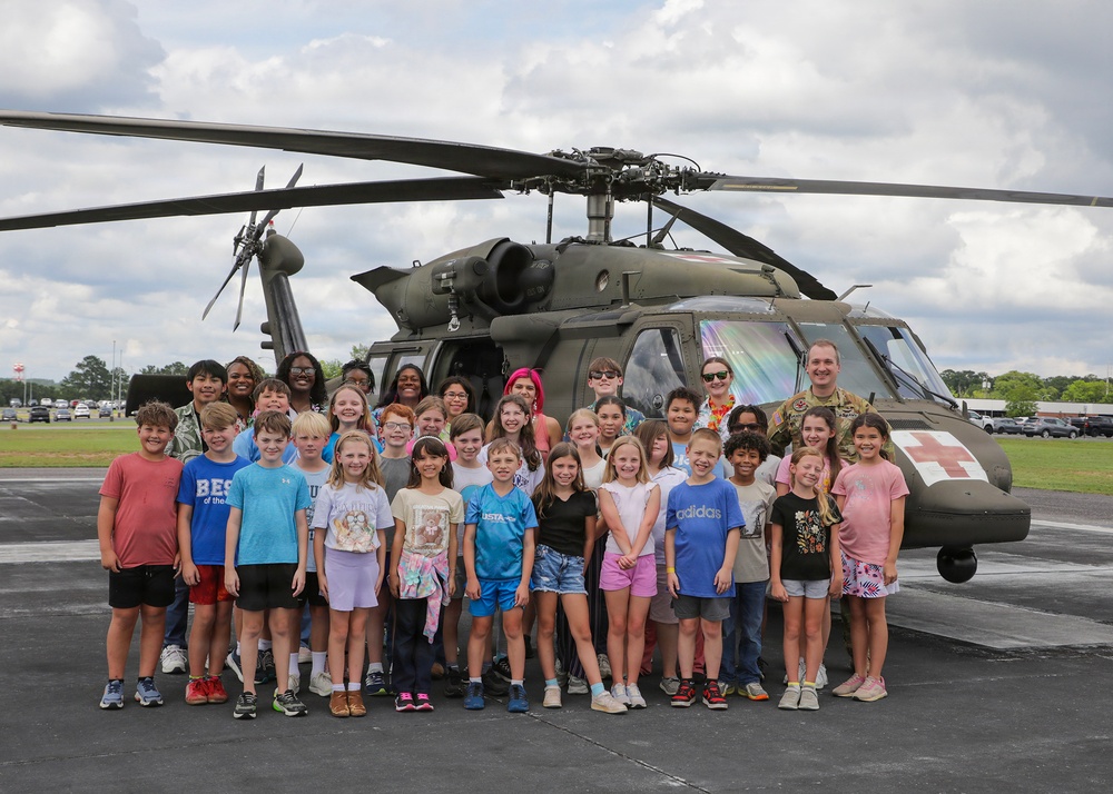 A large group of elementary school-aged children and several adults pose in front of a military UH-60 Black Hawk helicopter marked with a Red Cross emblem, indicating a medevac/medical variant. The group appears to be on a tarmac or airfield, suggesting an educational or community outreach event. At least one person in military camouflage uniform is visible among the adults.