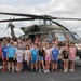 A large group of elementary school-aged children and several adults pose in front of a military UH-60 Black Hawk helicopter marked with a Red Cross emblem, indicating a medevac/medical variant. The group appears to be on a tarmac or airfield, suggesting an educational or community outreach event. At least one person in military camouflage uniform is visible among the adults.