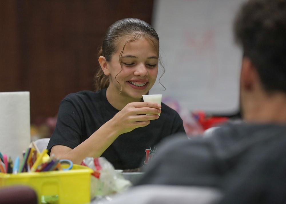 A young girl with braces and dark hair pulled back is smiling and holding a small white cup, appearing to be in a classroom or activity room setting. She is wearing a black t-shirt and seems engaged in a hands-on activity at a table. Another person is partially visible in the foreground with their back to the camera.