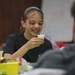 A young girl with braces and dark hair pulled back is smiling and holding a small white cup, appearing to be in a classroom or activity room setting. She is wearing a black t-shirt and seems engaged in a hands-on activity at a table. Another person is partially visible in the foreground with their back to the camera.