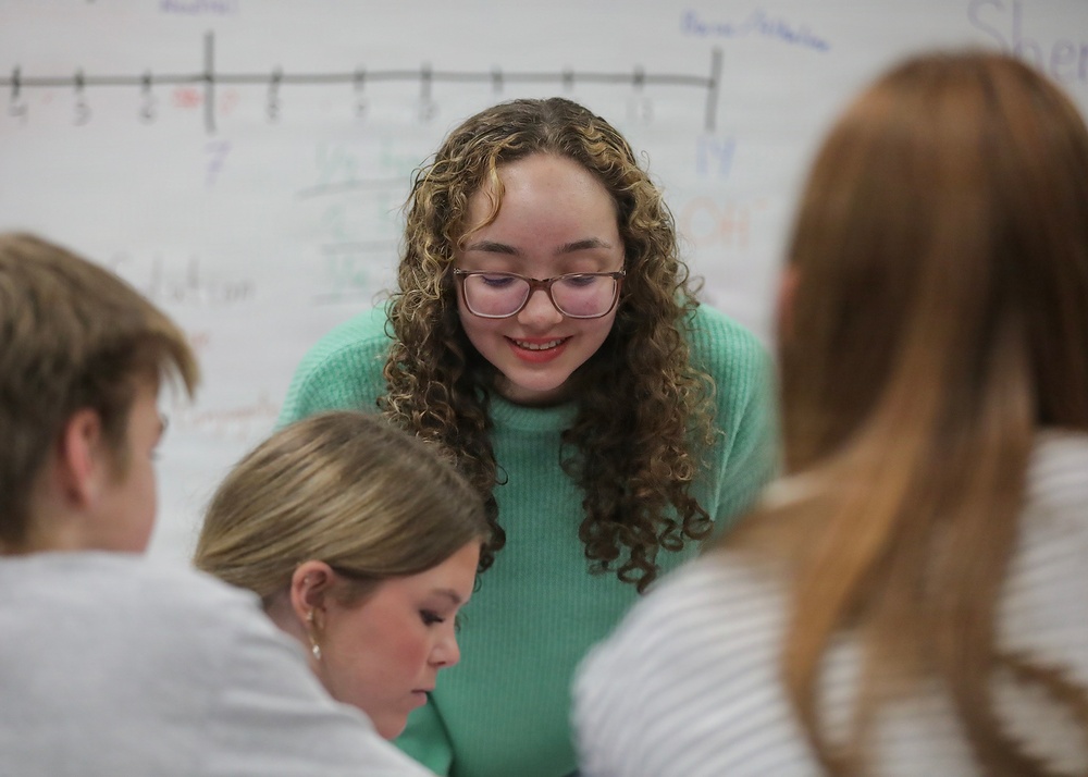 A young woman with curly hair and glasses wearing a teal/mint green sweater leans forward, smiling, while engaging with a small group of students in what appears to be a classroom setting. A whiteboard is visible in the background displaying a number line and various mathematical notations. The scene suggests a collaborative learning or tutoring interaction.