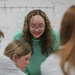 A young woman with curly hair and glasses wearing a teal/mint green sweater leans forward, smiling, while engaging with a small group of students in what appears to be a classroom setting. A whiteboard is visible in the background displaying a number line and various mathematical notations. The scene suggests a collaborative learning or tutoring interaction.