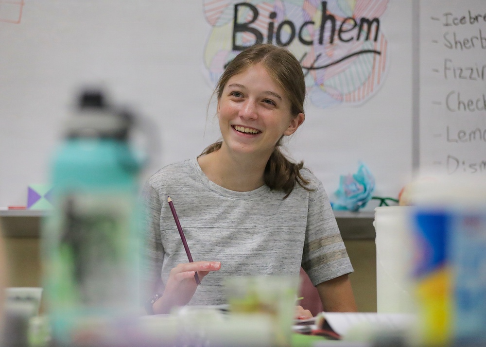 A young student is sitting at a classroom desk, smiling broadly while holding a pencil. She appears engaged and happy, likely participating in a biochemistry class as indicated by the 'Biochem' poster visible on the whiteboard behind her. The classroom setting includes various items on desks and a whiteboard with a partial list written on it.