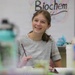 A young student is sitting at a classroom desk, smiling broadly while holding a pencil. She appears engaged and happy, likely participating in a biochemistry class as indicated by the 'Biochem' poster visible on the whiteboard behind her. The classroom setting includes various items on desks and a whiteboard with a partial list written on it.