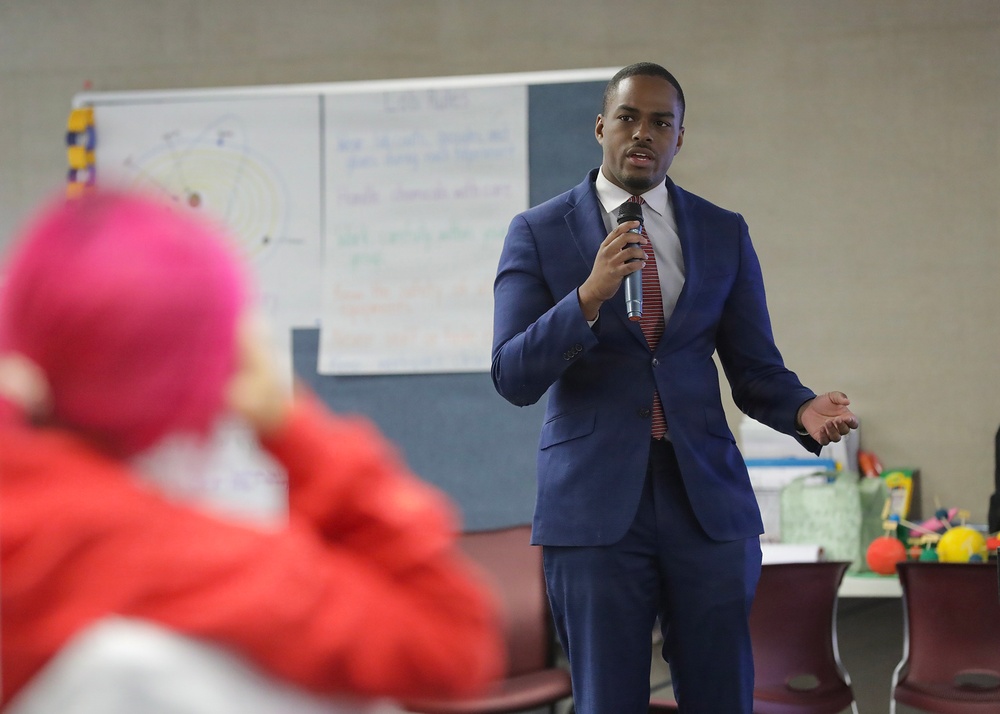 A man in a navy blue suit is speaking into a microphone in what appears to be a classroom or community meeting room. He is addressing an audience, with at least one attendee visible in the foreground wearing a bright pink head covering and red clothing. Behind him are whiteboards or flip charts with handwritten notes.