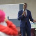 A man in a navy blue suit is speaking into a microphone in what appears to be a classroom or community meeting room. He is addressing an audience, with at least one attendee visible in the foreground wearing a bright pink head covering and red clothing. Behind him are whiteboards or flip charts with handwritten notes.