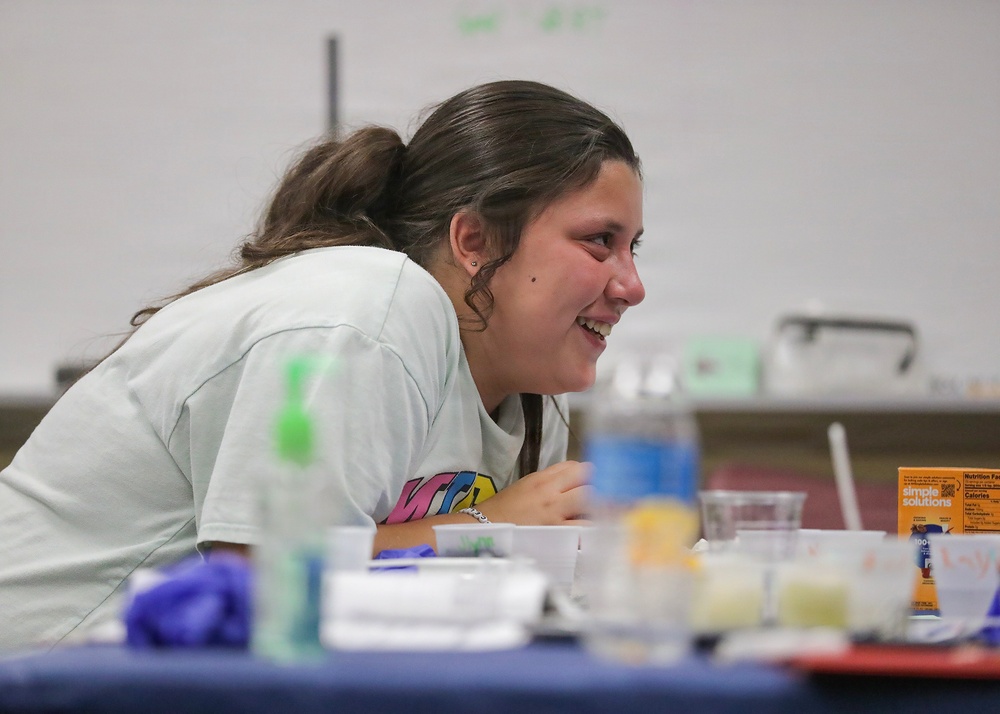 A young female student is smiling and laughing while leaning over a table covered with various lab materials and food/beverage items. The setting appears to be a classroom or educational workshop environment, with a whiteboard visible in the background. She appears engaged and enjoying the activity taking place.