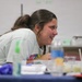 A young female student is smiling and laughing while leaning over a table covered with various lab materials and food/beverage items. The setting appears to be a classroom or educational workshop environment, with a whiteboard visible in the background. She appears engaged and enjoying the activity taking place.