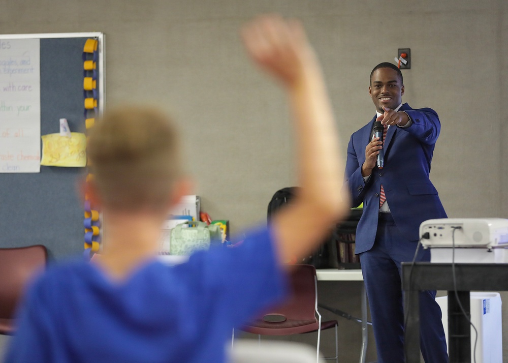 A well-dressed young man in a blue suit is speaking to a classroom audience while holding a microphone and pointing toward a student. In the foreground, a student wearing a blue hoodie has their hand raised. The setting appears to be a school classroom with educational materials visible on the walls.