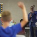 A well-dressed young man in a blue suit is speaking to a classroom audience while holding a microphone and pointing toward a student. In the foreground, a student wearing a blue hoodie has their hand raised. The setting appears to be a school classroom with educational materials visible on the walls.