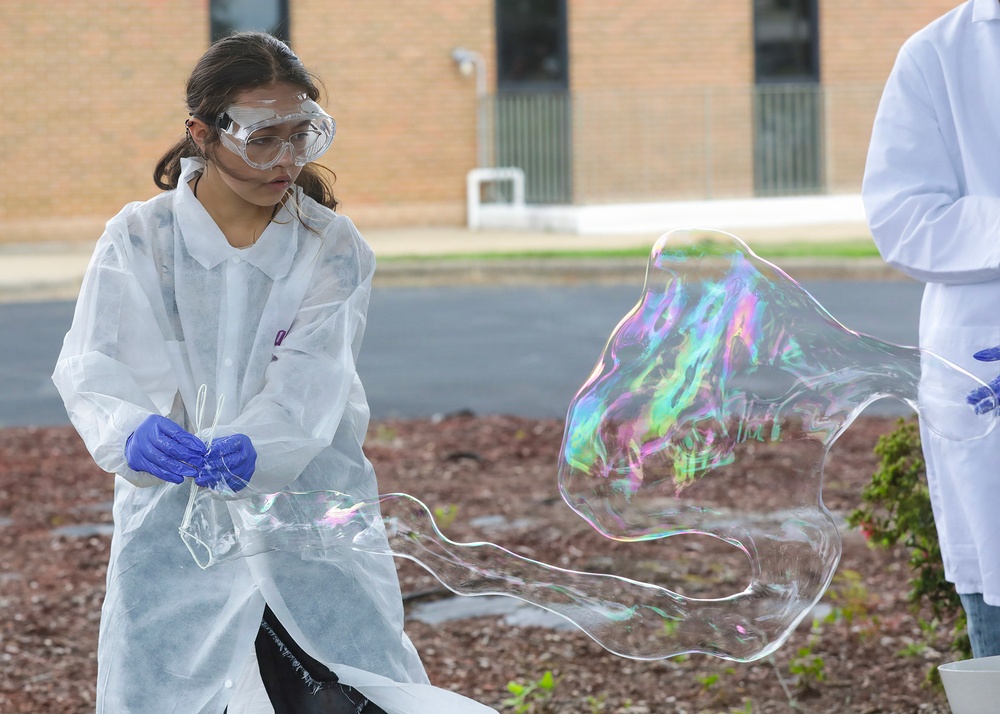 A young girl wearing a white lab coat, safety goggles, and blue latex gloves is creating a large soap bubble film outdoors, likely during a science demonstration or educational event. The iridescent bubble film displays vivid rainbow colors and is stretched into a large, irregular shape. A second person in a white lab coat is partially visible on the right side of the frame.
