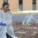 A young girl wearing a white lab coat, safety goggles, and blue latex gloves is creating a large soap bubble film outdoors, likely during a science demonstration or educational event. The iridescent bubble film displays vivid rainbow colors and is stretched into a large, irregular shape. A second person in a white lab coat is partially visible on the right side of the frame.