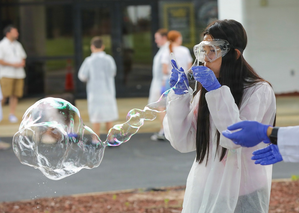 A young woman wearing a white lab coat, safety goggles, and blue latex gloves is blowing a large soap bubble outdoors. She appears to be participating in a science demonstration or educational activity, surrounded by other individuals similarly dressed in white lab coats. A large, iridescent soap bubble is visibly floating in front of her.