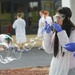 A young woman wearing a white lab coat, safety goggles, and blue latex gloves is blowing a large soap bubble outdoors. She appears to be participating in a science demonstration or educational activity, surrounded by other individuals similarly dressed in white lab coats. A large, iridescent soap bubble is visibly floating in front of her.