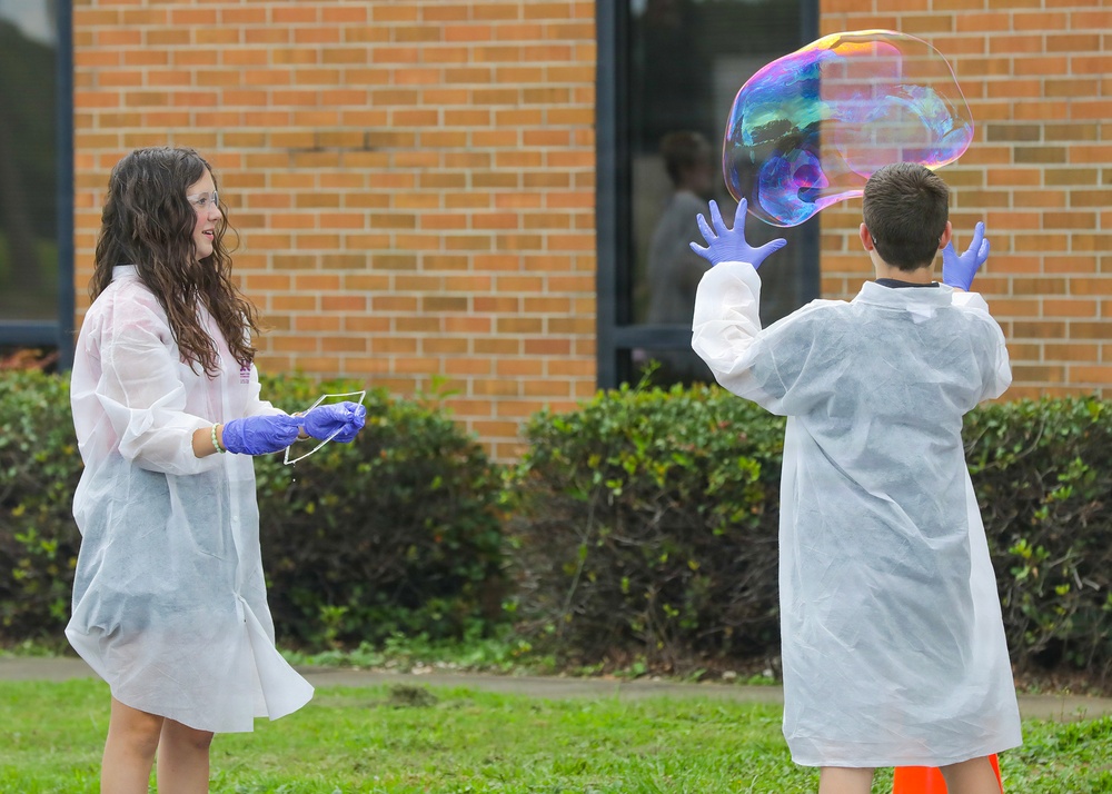 Two children wearing white lab coats and purple latex gloves are conducting an outdoor science experiment involving a giant soap bubble. One child faces the camera holding what appears to be a bubble wand or frame, while the other has their back turned with hands raised toward a large, iridescent bubble floating in the air. The activity appears to take place on a grassy area in front of a brick building, likely a school.