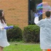 Two children wearing white lab coats and purple latex gloves are conducting an outdoor science experiment involving a giant soap bubble. One child faces the camera holding what appears to be a bubble wand or frame, while the other has their back turned with hands raised toward a large, iridescent bubble floating in the air. The activity appears to take place on a grassy area in front of a brick building, likely a school.