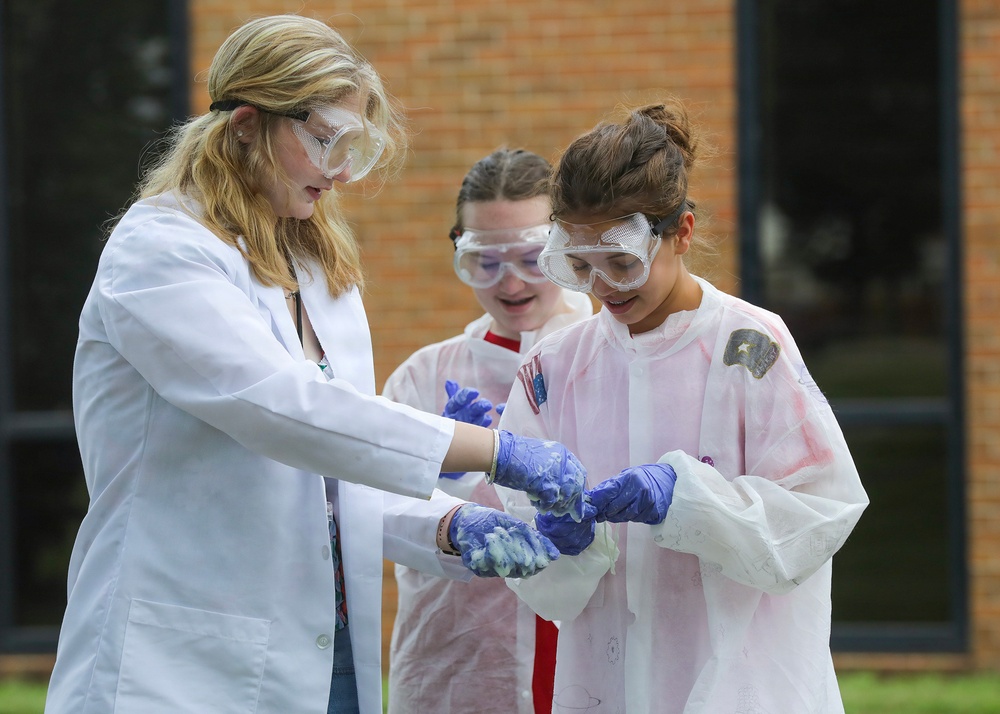 Three individuals are engaged in an outdoor science experiment or demonstration, wearing protective lab coats and safety goggles. Two younger students and what appears to be an older instructor or counselor are handling a blue, foamy substance with purple nitrile gloves. The setting appears to be outside a brick building, suggesting a school or educational camp environment.
