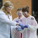 Three individuals are engaged in an outdoor science experiment or demonstration, wearing protective lab coats and safety goggles. Two younger students and what appears to be an older instructor or counselor are handling a blue, foamy substance with purple nitrile gloves. The setting appears to be outside a brick building, suggesting a school or educational camp environment.