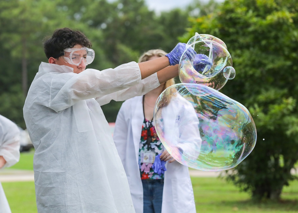 A young person wearing a white disposable lab coat, safety goggles, and blue latex gloves is manipulating large iridescent soap bubbles outdoors. A second person in a white lab coat stands in the background observing. The setting appears to be an outdoor science demonstration or educational event.