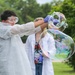 A young person wearing a white disposable lab coat, safety goggles, and blue latex gloves is manipulating large iridescent soap bubbles outdoors. A second person in a white lab coat stands in the background observing. The setting appears to be an outdoor science demonstration or educational event.
