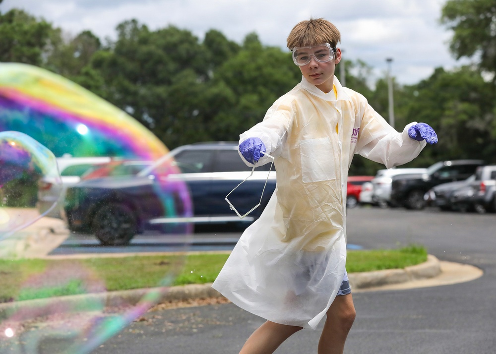 A young person wearing a white disposable lab coat, safety goggles, and blue nitrile gloves stands in a parking lot surrounded by large iridescent soap bubbles. They are holding what appears to be a face mask in one hand and appear to be engaged in a science demonstration or outdoor experiment. The setting includes a parking lot with vehicles and trees in the background under a partly cloudy sky.