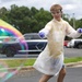 A young person wearing a white disposable lab coat, safety goggles, and blue nitrile gloves stands in a parking lot surrounded by large iridescent soap bubbles. They are holding what appears to be a face mask in one hand and appear to be engaged in a science demonstration or outdoor experiment. The setting includes a parking lot with vehicles and trees in the background under a partly cloudy sky.