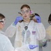 A group of young students participate in a hands-on science education program, wearing protective lab coats, safety goggles, and purple nitrile gloves. The central student is blowing soap bubbles using a geometric wire frame, with several bubbles visible floating in the air. The lab coats display the 'OEOP' (Office of Educational Outreach Programs) logo with the website 'usieop.com'.