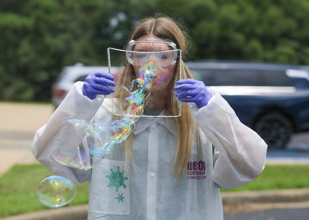 A person wearing a white lab coat, safety goggles, and purple nitrile gloves is blowing soap bubbles through a square wire frame outdoors. The lab coat displays a logo and text reading 'usaeop.com', suggesting this is part of a science educational outreach demonstration. Multiple iridescent soap bubbles are visible in various stages of formation.