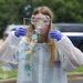 A person wearing a white lab coat, safety goggles, and purple nitrile gloves is blowing soap bubbles through a square wire frame outdoors. The lab coat displays a logo and text reading 'usaeop.com', suggesting this is part of a science educational outreach demonstration. Multiple iridescent soap bubbles are visible in various stages of formation.