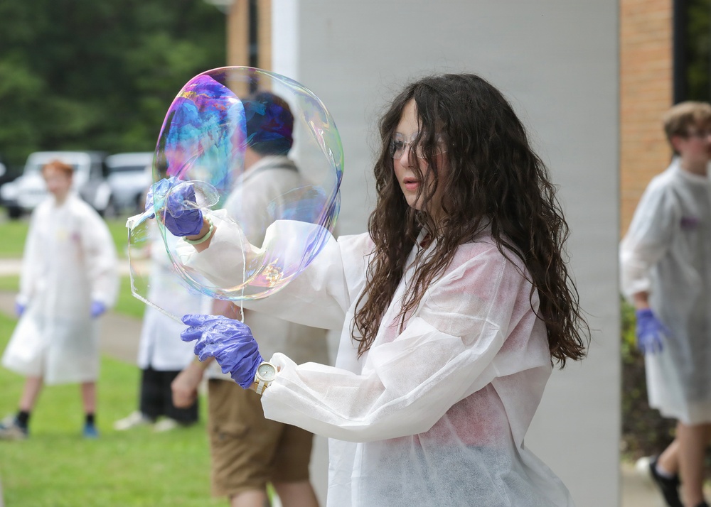 A person wearing a white disposable lab coat, blue nitrile gloves, safety goggles, and a wristwatch is manipulating a large, colorful soap bubble outdoors. The bubble displays vivid iridescent colors including blue, purple, and pink. In the background, other individuals in similar white protective gear are visible, suggesting a group science demonstration or educational activity.